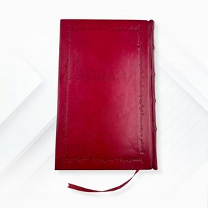 Puede incluir: Un diario rojo encuadernado en cuero con un borde decorativo y un marcador de cinta a juego. La cubierta del diario es de color rojo intenso y los bordes de las p&aacute;ginas son visibles. El dise&ntilde;o general es cl&aacute;sico y elegante.