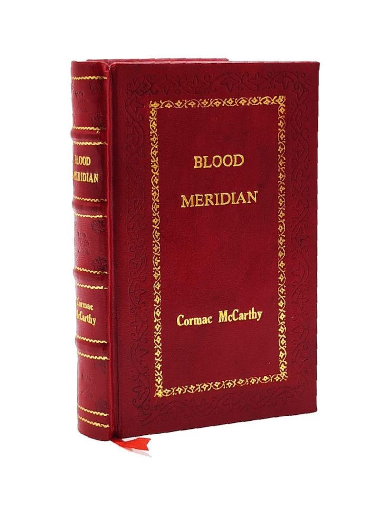May include: A hardcover book titled "Blood Meridian" by Cormac McCarthy. The book has a deep red cover with gold lettering and a decorative gold border. The spine also features gold lettering. A red ribbon bookmark is visible.