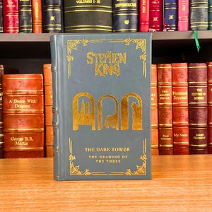 K&ouml;nnte beinhalten: Ein Hardcover-Buch mit dem Titel "The Dark Tower: The Drawing of the Three" von Stephen King. Das Buch hat einen grauen Einband mit goldfarbenen Buchstaben und Illustrationen. Das Buch wird vor einem B&uuml;cherregal mit anderen B&uuml;chern pr&auml;sentiert.