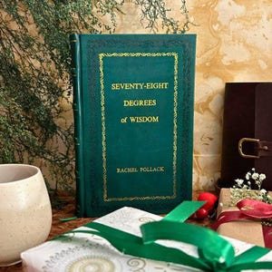 Op de afbeelding: Een donkergroen hardcover boek met gouden letters en een decoratieve rand. Het boek heet "SEVENTY-EIGHT DEGREES of WISDOM" van Rachel Pollack. Een ingepakt cadeau met een groen lint staat op de voorgrond.