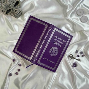 Può includere: Un libro viola aperto, intitolato "The Lesser Key of Solomon Goetia", con dettagli argentati e un segnalibro. Una chiave decorativa, pietre di ametista e un teschio sono disposti su una superficie di raso.