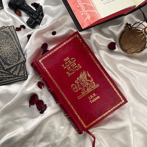 Puede incluir: Una edici&oacute;n en cuero rojo de "El Se&ntilde;or de los Anillos" de J.R.R. Tolkien, con letras doradas y un emblema decorativo. El libro est&aacute; abierto en una p&aacute;gina con detalles en rojo. Otros elementos incluyen cartas del tarot, un objeto decorativo negro y una jaula dorada.