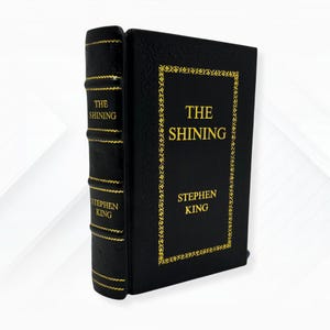 Könnte beinhalten: Ein schwarzes Hardcover-Buch mit dem Titel "THE SHINING" von Stephen King in goldener Schrift. Der Einband hat einen goldenen Zierrand. Der Buchrücken zeigt ebenfalls Titel und Autor in Gold.