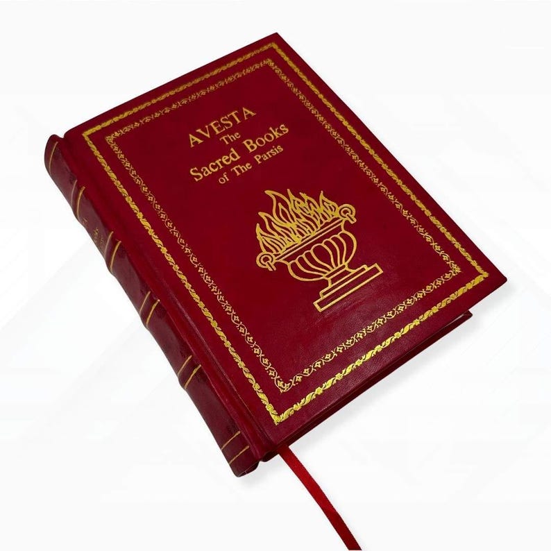 Op de afbeelding: Een rood hardcover boek getiteld "AVESTA The Sacred Books of The Parsis" met gouden letters en een decoratieve gouden rand. De omslag heeft een gouden illustratie van een brandende urn. Een rood lint-bladwijzer is zichtbaar.
