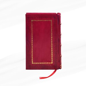 Op de afbeelding: Een dieprood hardcover boek met gouden decoratieve accenten en een bijpassend rood lint als bladwijzer. De kaft heeft een gouden rechthoekige rand met sierlijke details. De rug heeft gouden accenten.