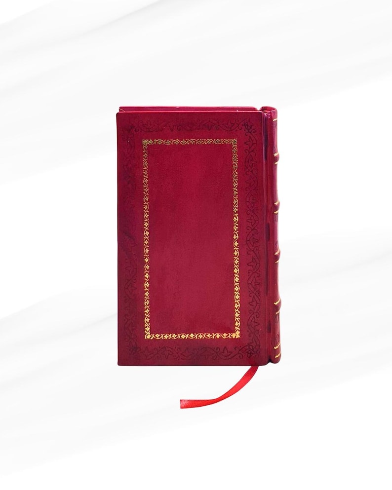 Op de afbeelding: Een dieprood hardcover boek met gouden decoratieve accenten en een bijpassend rood lint als bladwijzer. De kaft heeft een gouden rechthoekige rand met sierlijke details. De rug heeft gouden accenten.
