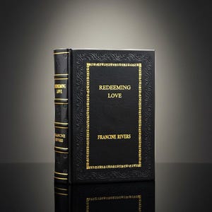 May include: A black hardcover book titled "Redeeming Love" by Francine Rivers, with gold lettering and a decorative gold border. The spine also has gold lettering. The book is reflected in a glossy surface. The book's theme is love.