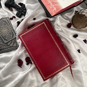 Puede incluir: Un libro rojo encuadernado en cuero con detalles de filigrana dorada y un marcador a juego. El libro est&aacute; sobre una superficie de sat&eacute;n blanco, rodeado de cartas del tarot, p&eacute;talos de rosa secos y otros objetos decorativos.