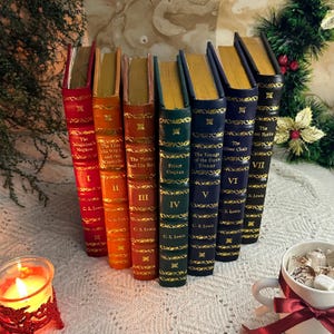 Op de afbeelding: Een verzameling van zeven hardcover boeken met goudkleurige pagina's en decoratieve gouden accenten. De ruggen zijn in verschillende kleuren, waaronder rood, oranje, groen en blauw. Titels en auteursnamen zijn zichtbaar. Een brandende kaars en een mok marshmallows staan op de achtergrond.