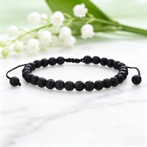Peut inclure: Bracelet de perles en pierre de lave noire. Le bracelet est composé de perles noires rondes et poreuses sur un cordon noir. Il est réglable grâce à un nœud coulissant. Des fleurs blanches et des feuilles vertes sont en arrière-plan.