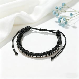 Op de afbeelding: Zwarte leren armband met zilverkleurige cilindrische en bolvormige kralen. De armband heeft verstelbare zwarte koorden. De sieraden worden gepresenteerd op een witte ondergrond met zachte stof en kleine blauwe bloemen.