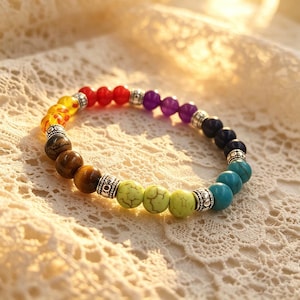 May include: A multi-coloured beaded bracelet with a range of hues, including red, yellow, green, and blue. The bracelet is made with different types of beads and silver-toned spacers. The bracelet is displayed on a white lace surface.