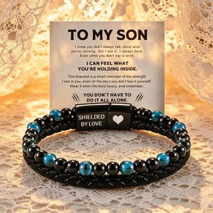 Könnte beinhalten: Ein schwarzes geflochtenes Lederarmband mit schwarzen und blauen Perlen. Das Armband hat einen Magnetverschluss mit den Worten "SHIELDED BY LOVE" und einem Herzsymbol. Eine Karte hinter dem Armband lautet "TO MY SON".