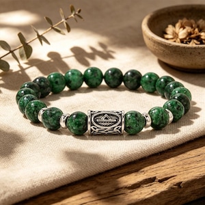 May include: A bracelet with dark green, spherical beads and a silver-coloured cylindrical centrepiece with an engraved design. The bracelet is displayed on a neutral-coloured cloth.