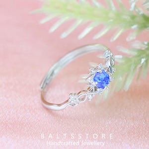 Peut inclure: Bague en argent ornée d'une pierre bleue ronde centrale, entourée de petits brillants. La bague présente un design ajouré avec des motifs de filigrane. Le texte "BALTSSTORE Handcrafted Jewellery" est visible.