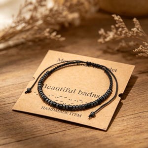 May include: A black beaded bracelet with the text "Beautiful badas" on a brown card. The bracelet is made of small, dark gray beads strung on a black cord. The card also says "HANDMADE ITEM".