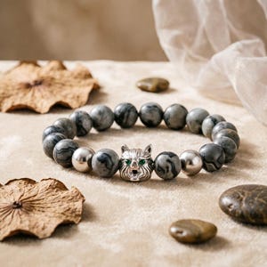 May include: A beaded bracelet featuring gray and silver beads. The focal point is a silver cat head with green eyes. The bracelet is displayed on a neutral-toned surface with decorative elements.