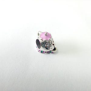 May include: A silver-coloured charm in the shape of a teapot. The teapot has a pink lid with white dots and a pink rim. The base of the teapot has green and purple accents. A small clear gem is set on the front of the teapot.
