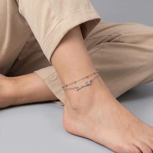 May include: A silver-colored double-layered anklet with the name "Camila" in cursive script. The anklet features small, round beads and is worn on the ankle. The background is a neutral gray.