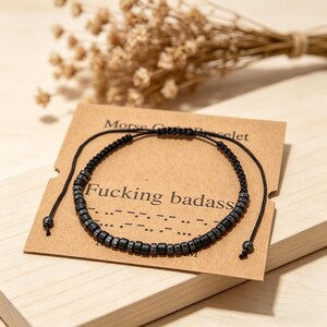 Op de afbeelding: Zwart kralenarmband met de tekst "Fucking badass" op een bruine kaart. De armband is gemaakt van kleine, donkergrijze kralen en zwart koord. De kaart heeft ook de tekst "Morse Code Bracelet".