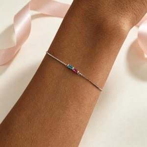May include: A silver bracelet with a fine chain, adorned with two rectangular gemstones, one blue and one pink. The bracelet is displayed on a wrist, highlighting the jewellery's design. The bracelet is a piece of jewellery.