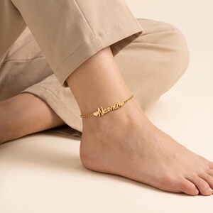 May include: A gold-colored anklet with the name "Naomi" in cursive script. The anklet features small heart-shaped charms on either side of the name. The anklet is worn on an ankle.