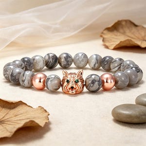 May include: A beaded bracelet featuring gray and white marbled stone beads, with two rose gold beads and a rose gold wolf head charm with green eyes. The bracelet is displayed on a neutral surface.