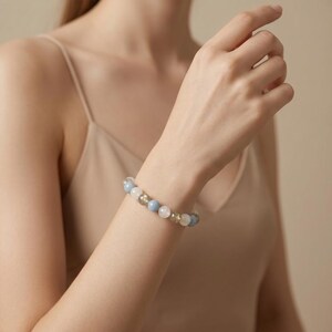 May include: A beaded bracelet featuring light blue, white, and gold-colored spherical beads. The bracelet is worn on a wrist, with the beads appearing to be approximately 0.5 inches in diameter. The bracelet is a piece of jewelry.