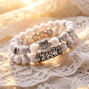 May include: Two beaded bracelets with white and grey marble-like beads. A silver-coloured owl charm and decorative metal accents are featured on one bracelet. The bracelets are displayed on a white lace surface.