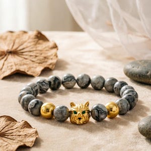 May include: A beaded bracelet featuring gray and black marbled beads, gold-colored spheres, and a gold-colored cat head charm with green eyes. The bracelet is displayed on a neutral surface.