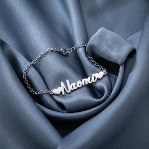 Könnte beinhalten: Ein silbernes Armband mit dem Namen "Naomi" in Schreibschrift und kleinen Herzanhängern. Das Armband liegt auf einem blauen Satin-Stoff, der einen luxuriösen Hintergrund bildet.