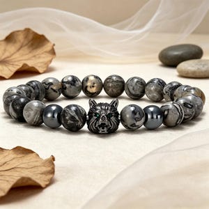 May include: A beaded bracelet featuring a silver wolf head with green eyes. The bracelet is composed of gray and black patterned stone beads and two dark gray metallic beads. The bracelet is displayed on a neutral background.