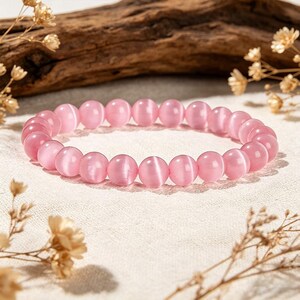 May include: A pink beaded bracelet. The bracelet features round, polished beads in a soft pink colour. The beads are strung together, forming a circular shape. The bracelet is displayed on a white surface with dried flowers and a piece of wood in the background.
