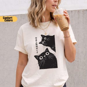 Pu&ograve; includere: T-shirt color crema con grafica di due gatti neri con grandi occhi. Testo giapponese stampato verticalmente sul lato sinistro. Parte della collezione Comfort Colors.