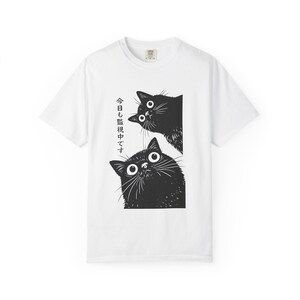 Pu&ograve; includere: T-shirt bianca con un'illustrazione in bianco e nero di due gatti. I gatti sbirciano oltre un bordo e hanno grandi occhi e baffi. Il testo giapponese &egrave; stampato verticalmente sul lato sinistro del disegno.