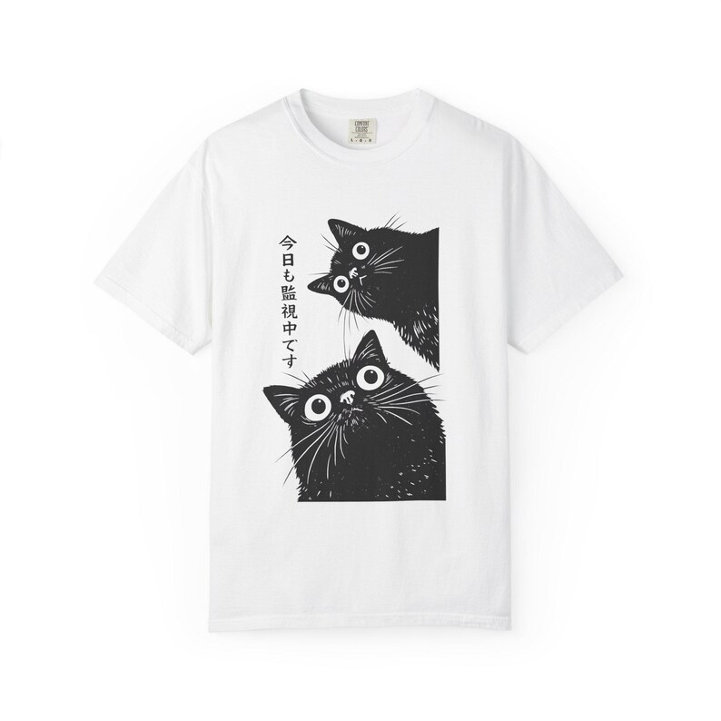 Pu&ograve; includere: T-shirt bianca con un'illustrazione in bianco e nero di due gatti. I gatti sbirciano oltre un bordo e hanno grandi occhi e baffi. Il testo giapponese &egrave; stampato verticalmente sul lato sinistro del disegno.