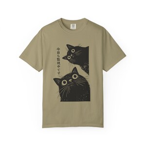 Pu&ograve; includere: Una maglietta color cachi con una grafica di due gatti neri con grandi occhi. I gatti guardano in alto e il testo giapponese &egrave; stampato verticalmente a sinistra. La maglietta ha maniche corte e girocollo.
