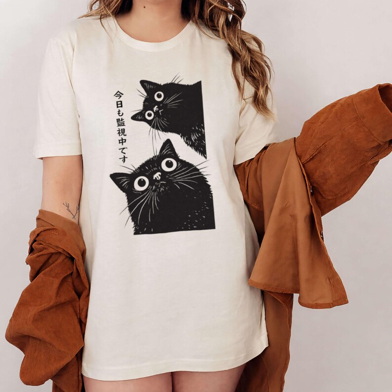 Pu&ograve; includere: T-shirt color crema con grafica di due gatti neri con grandi occhi spalancati. Testo giapponese stampato sul lato sinistro. I gatti spuntano da una cornice rettangolare. Una giacca marrone &egrave; drappeggiata sul braccio.