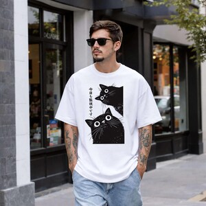 Pu&ograve; includere: T-shirt bianca con una grafica di due gatti neri con grandi occhi. Il testo giapponese sulla maglietta recita: "Ti stiamo guardando oggi". La persona indossa anche occhiali da sole e jeans.