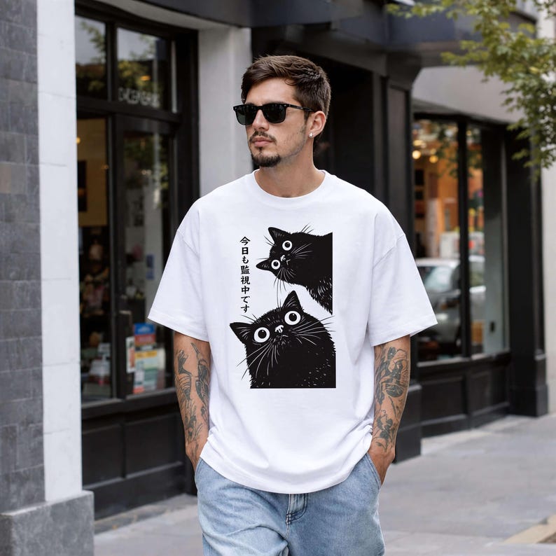 Pu&ograve; includere: T-shirt bianca con una grafica di due gatti neri con grandi occhi. Il testo giapponese sulla maglietta recita: "Ti stiamo guardando oggi". La persona indossa anche occhiali da sole e jeans.