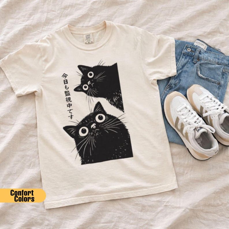 Pu&ograve; includere: T-shirt color crema con grafica di due gatti neri con grandi occhi e baffi. Testo giapponese stampato sulla maglietta. La maglietta &egrave; abbinata a jeans blu e scarpe da ginnastica bianche.