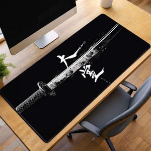 May include: A rectangular black desk mat featuring a white graphic illustration of a katana sword positioned diagonally. The design includes white Japanese kanji characters and a distressed, ink-splatter effect along the blade of the sword.