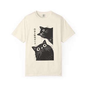Pu&ograve; includere: T-shirt color crema con un'illustrazione nera di due gatti che sbucano da un bordo rettangolare. I gatti hanno grandi occhi spalancati. Testo giapponese stampato verticalmente a sinistra dei gatti.