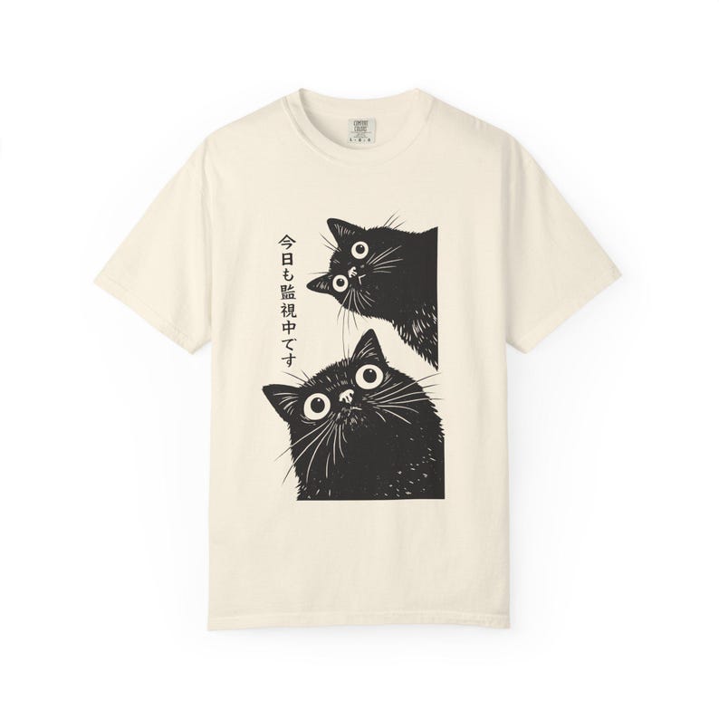 Pu&ograve; includere: T-shirt color crema con un'illustrazione nera di due gatti che sbucano da un bordo rettangolare. I gatti hanno grandi occhi spalancati. Testo giapponese stampato verticalmente a sinistra dei gatti.