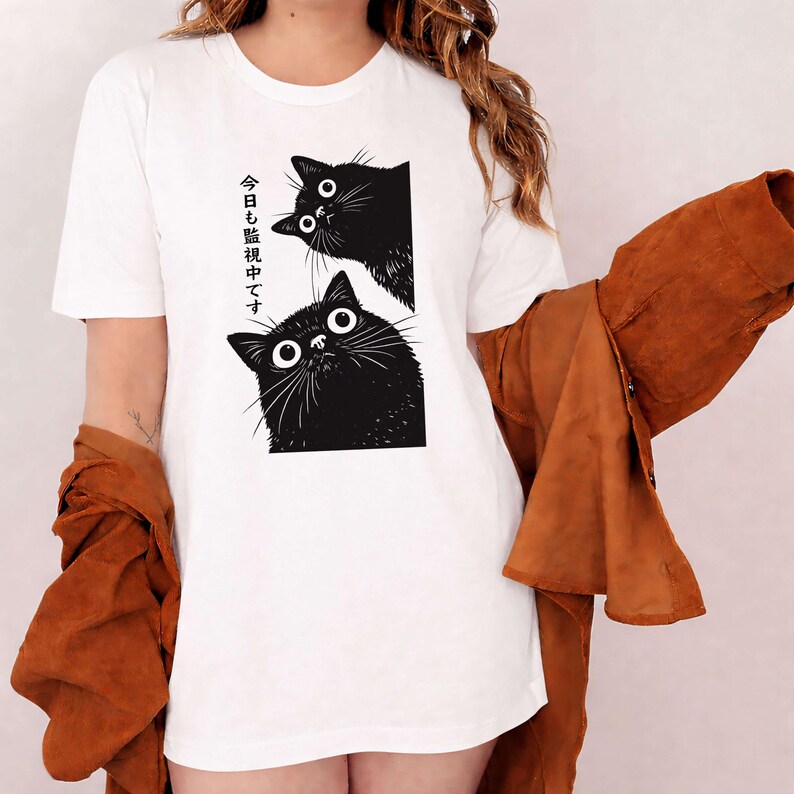 Pu&ograve; includere: T-shirt bianca con un'illustrazione in bianco e nero di due gatti con grandi occhi. Il testo giapponese sulla maglietta recita "Ti stiamo guardando oggi."