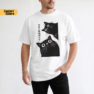 Pu&ograve; includere: T-shirt bianca con un'illustrazione in bianco e nero di due gatti con grandi occhi. Il testo giapponese sulla maglietta recita "Stiamo guardando anche oggi". La maglietta ha una vestibilit&agrave; ampia.