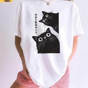 Pu&ograve; includere: T-shirt bianca con grafica nera di due gatti con grandi occhi. Il testo giapponese sulla maglietta recita "Ti stiamo guardando oggi". La maglietta fa parte della collezione Comfort Colors.