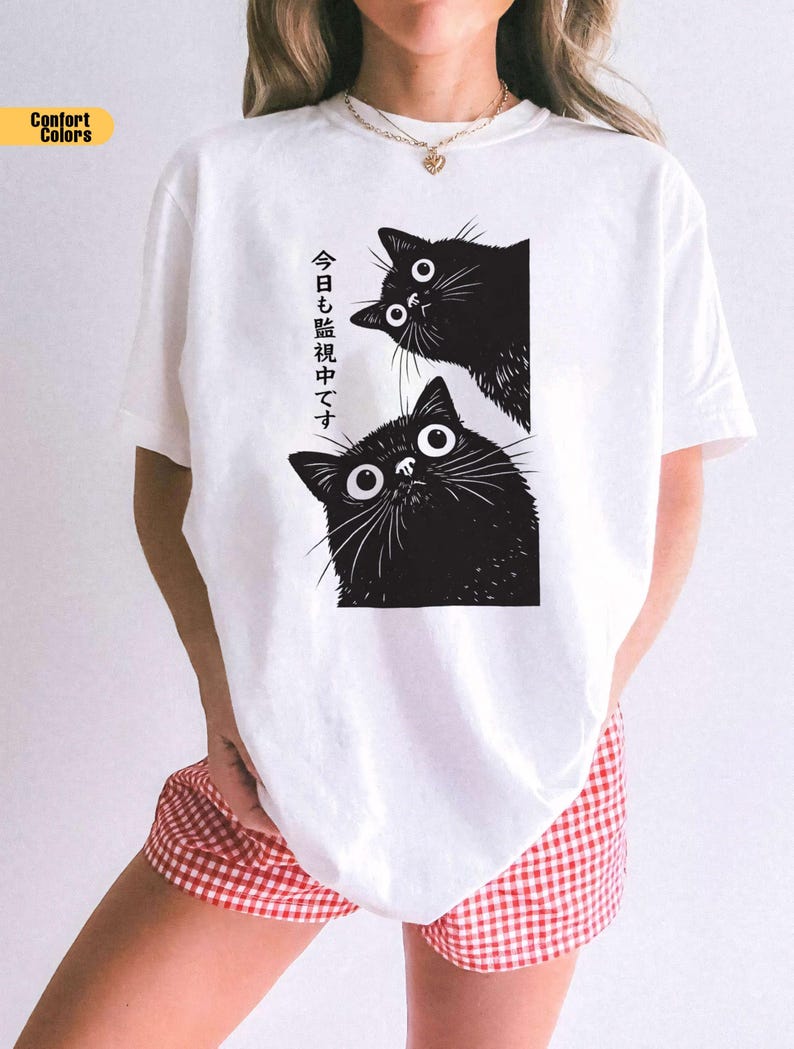Pu&ograve; includere: T-shirt bianca con grafica nera di due gatti con grandi occhi. Il testo giapponese sulla maglietta recita "Ti stiamo guardando oggi". La maglietta fa parte della collezione Comfort Colors.