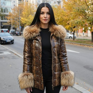 Pode incluir: Casaco castanho com padrão de pele de cobra com gola e punhos de pele. O casaco é fechado com zíper e usado sobre um top preto. O fundo mostra uma rua da cidade com carros e árvores.