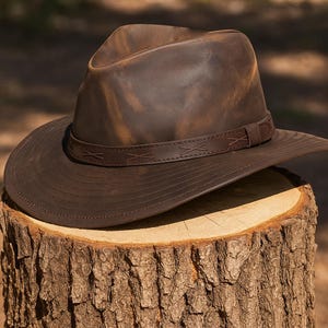 Puede incluir: Un sombrero de cuero marrón con ala ancha y una banda alrededor de la corona. El sombrero tiene un acabado envejecido, lo que le da un aspecto robusto y vintage. El sombrero está sobre un tocón.